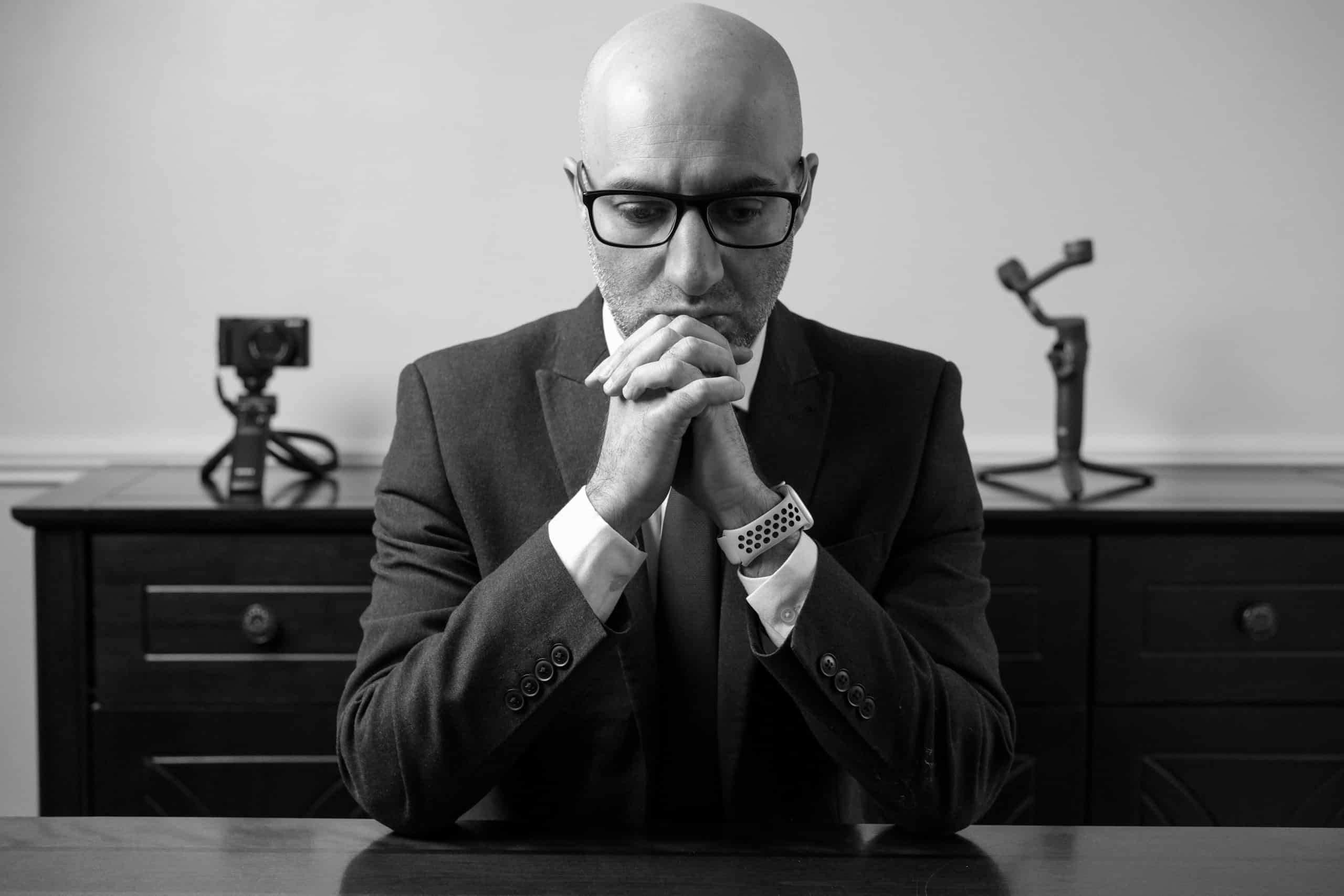 An executive in a dark suit seated at a desk, hands clasped beneath his chin in a posture of deep deliberation.