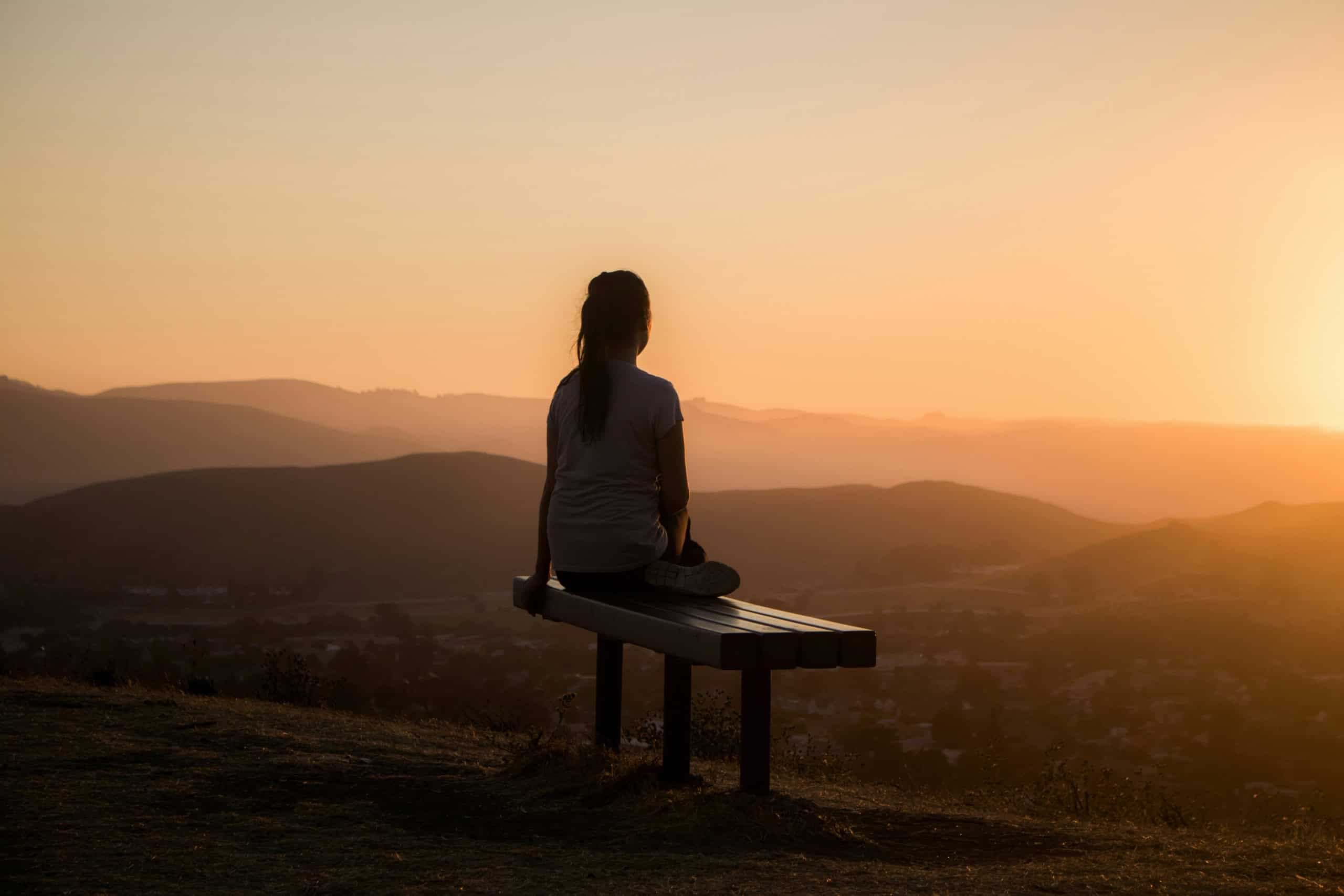 A person sits alone on a wooden bench atop a hill at golden hour, gazing out across layered mountains and a warm, expansive sky.